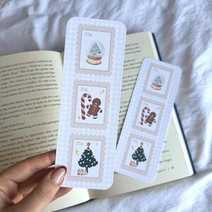 May include: Two Christmas-themed bookmarks with postage stamp designs. Each bookmark features three stamps with festive illustrations: a snow globe, a gingerbread man, and a Christmas tree. The stamps are framed with a light pink border.