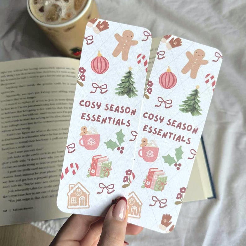 May include: Two white bookmarks with a repeating pattern of gingerbread men, Christmas trees, ornaments, and other holiday-themed illustrations. The text "COSY SEASON ESSENTIALS" is printed on the bookmarks. A hand holds the bookmarks near a book and a cup of coffee.