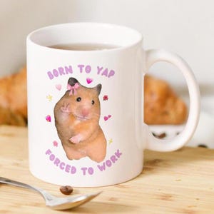 May include: White ceramic mug with a pink and purple design. The mug features a hamster with a pink bow and the text "Born to Yap Forced to Work".