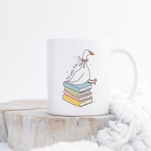 May include: White ceramic mug featuring a cartoon goose with a pink bow, perched atop a stack of colorful books. The mug has the text "Jolly Goose Book Club" in a whimsical font. The design is in soft pastel colors.