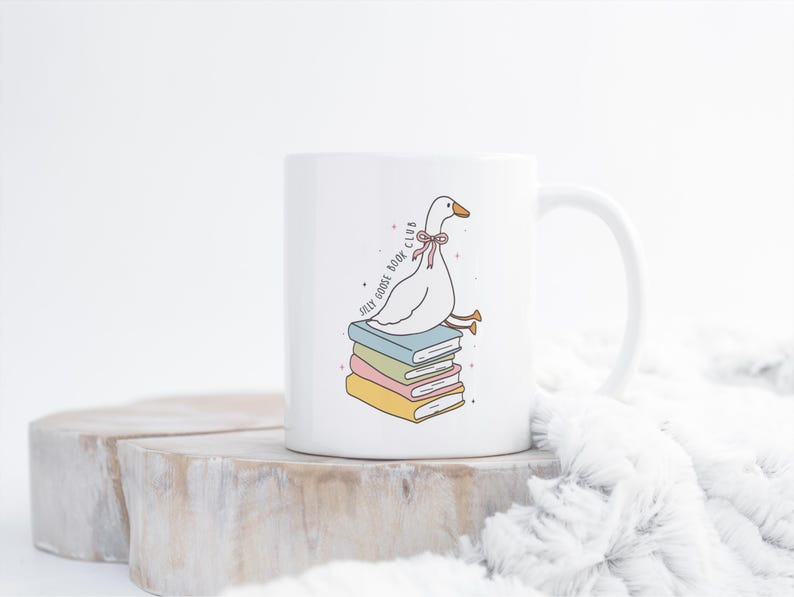 May include: White ceramic mug featuring a cartoon goose with a pink bow, perched atop a stack of colorful books. The mug has the text "Jolly Goose Book Club" in a whimsical font. The design is in soft pastel colors.