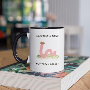May include: White ceramic mug with a black handle and rim, featuring a cartoon worm wearing a cowboy hat. The mug has the text "SOMETIMES I THINK BUT THEN I FORGET". The mug is sitting on a book.