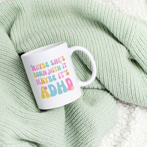 May include: White ceramic mug with the text "MAYBE SHE'S BORN WITH IT MAYBE IT'S ADHD" in colorful, retro-style lettering. The mug is set against a soft, light green knitted blanket.