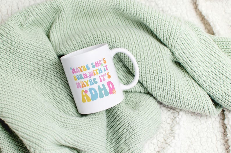 May include: White ceramic mug with the text "MAYBE SHE'S BORN WITH IT MAYBE IT'S ADHD" in colorful, retro-style lettering. The mug is set against a soft, light green knitted blanket.