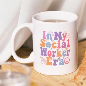 Könnte beinhalten: Weißer Keramikbecher mit dem Text "In My Social Worker Era" in farbenfrohen Buchstaben im Retro-Stil. Der Becher hat einen großen Henkel und ist mit einer hellen Flüssigkeit gefüllt.