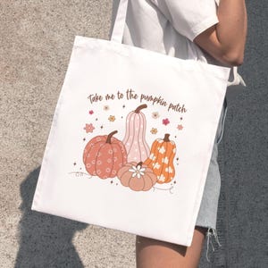 Può includere: Borsa tote bianca con un'illustrazione di zucche arancioni, rosa e marroni. Il testo "Take me to the pumpkin patch" è stampato sopra le zucche. La borsa è tenuta da una persona.
