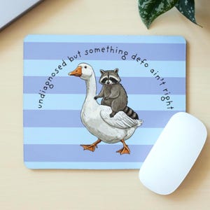 May include: A mouse mat with a blue and white striped background, featuring a cartoon goose with a raccoon riding on its back. The text "undiagnosed but something defo ain't right" is printed around the image. A white computer mouse is visible.