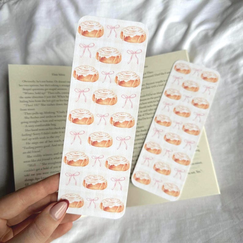 Pode incluir: Dois marcadores retangulares com um design em aquarela de rolinhos de canela e la&ccedil;os rosa. Os marcadores s&atilde;o segurados sobre um livro aberto. O design apresenta um padr&atilde;o repetido dos produtos de panifica&ccedil;&atilde;o e la&ccedil;os.
