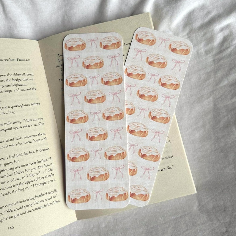 Pode incluir: Dois marcadores retangulares com um padr&atilde;o repetido de rolinhos de canela em aquarela e la&ccedil;os rosa. Os marcadores est&atilde;o colocados nas p&aacute;ginas de um livro aberto. Os marcadores s&atilde;o de cor creme claro.