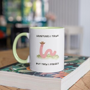 May include: White ceramic mug with a light green handle and interior. The mug features a cartoon worm wearing a cowboy hat, with the text "SOMETIMES I THINK BUT THEN I FORGET". The mug is sitting on a book.