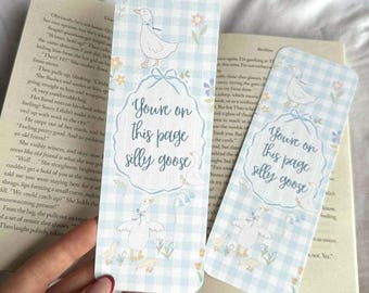 May include: Two rectangular bookmarks with a light blue gingham pattern and the text "You're on this page silly goose." Each bookmark features a white goose illustration and floral accents. The bookmarks are on top of an open book.