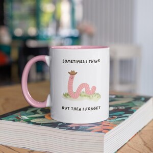 May include: White ceramic mug with a pink interior and handle. The mug features a cartoon worm wearing a cowboy hat, with the text "SOMETIMES I THINK BUT THEN I FORGET". The mug is sitting on a book.