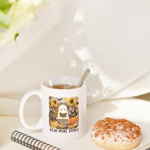 May include: White ceramic mug with a Halloween-themed illustration of a ghost reading a book, surrounded by pumpkins and sunflowers. The mug has the text "READ MORE BOOKS". A donut and a notebook are also in the image.