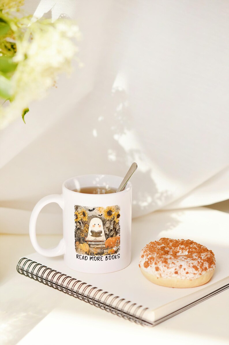 May include: White ceramic mug with a Halloween-themed illustration of a ghost reading a book, surrounded by pumpkins and sunflowers. The mug has the text "READ MORE BOOKS". A donut and a notebook are also in the image.