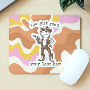 May include: A rectangular mouse mat with a retro-style design in pink, orange, and brown. The mat features a cartoon frog dressed as a cowboy, holding a gun, with the text "you just yee'd your last haw."