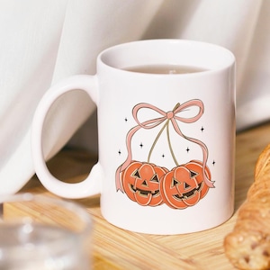 May include: White ceramic mug with a Halloween design. The design shows two orange jack-o'-lanterns with smiling faces, linked by a pink bow and ribbons. Small black stars are scattered around the design. The mug has a curved handle.