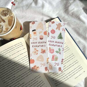 May include: Two decorative bookmarks with a cozy season theme. One features a coffee cup, pumpkin, and sweater. The other shows a gingerbread man, Christmas tree, and mug. Text on both reads "COSY SEASON ESSENTIALS."