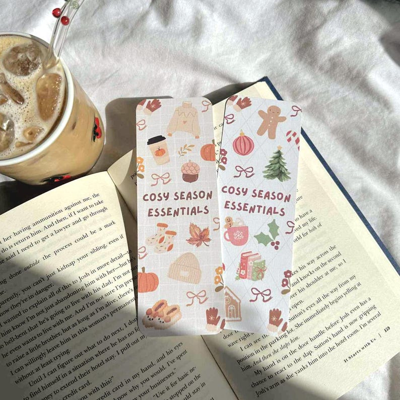 May include: Two decorative bookmarks with a cozy season theme. One features a coffee cup, pumpkin, and sweater. The other shows a gingerbread man, Christmas tree, and mug. Text on both reads "COSY SEASON ESSENTIALS."