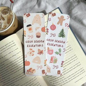 May include: Two decorative bookmarks with a cozy season theme. One features a sweater, coffee, and pumpkin, while the other shows a gingerbread man, tree, and mug. The text "COSY SEASON ESSENTIALS" is printed on both.