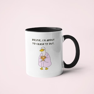May include: White ceramic mug with a black handle and rim. The mug features an illustration of a duck wearing a pink robe and holding a cup. Text on the mug reads, "Bestie, I'm about to crash TF out."
