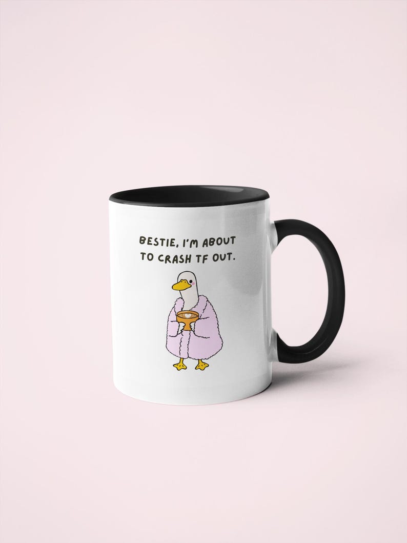 May include: White ceramic mug with a black handle and rim. The mug features an illustration of a duck wearing a pink robe and holding a cup. Text on the mug reads, "Bestie, I'm about to crash TF out."