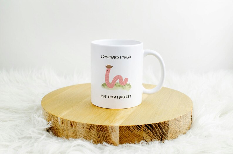 May include: White ceramic mug with a whimsical design featuring a pink worm wearing a cowboy hat, resting on a patch of green grass. The mug has the text "Sometimes I think, but then I forget". The mug is on a wooden stand.