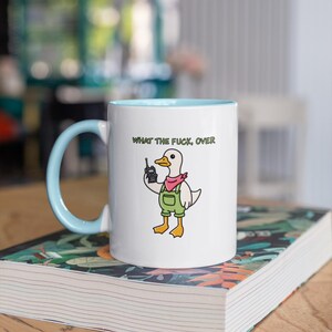 May include: White ceramic mug with a light blue interior and handle. The mug features a cartoon duck wearing overalls and a pink bandana, holding a cell phone. The text "WHAT THE FUCK, OVER" is printed above the duck.