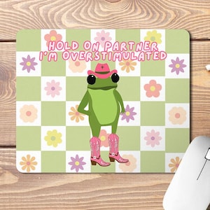 Op de afbeelding: Een groene kikker met een roze cowboyhoed en roze cowboylaarzen op een groen-wit geruite achtergrond. De tekst "HOLD ON PARTNER I'M OVERSTIMULATED" staat in roze letters boven de kikker.