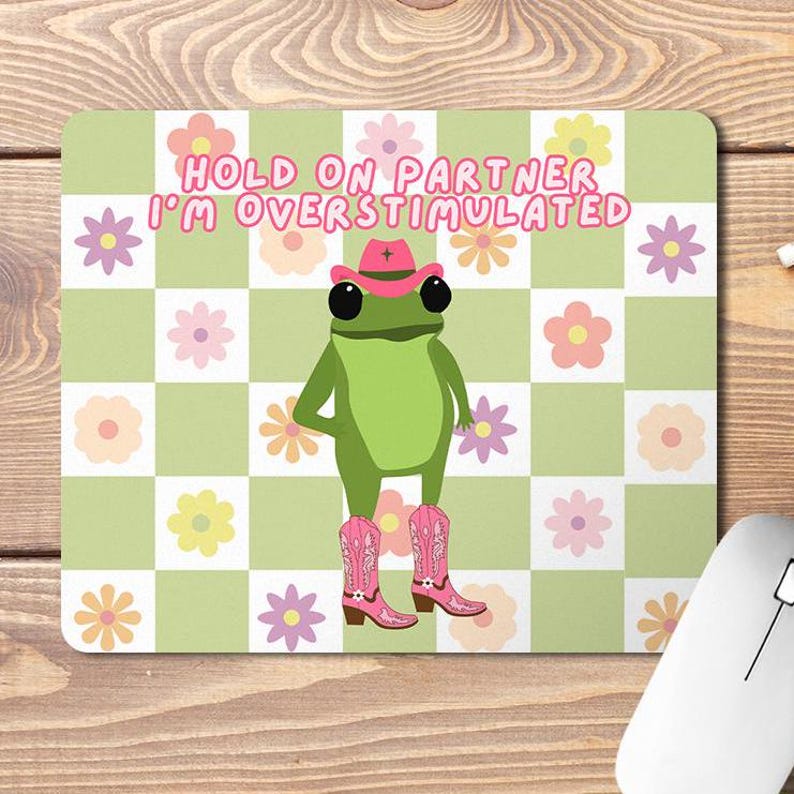 Op de afbeelding: Een groene kikker met een roze cowboyhoed en roze cowboylaarzen op een groen-wit geruite achtergrond. De tekst "HOLD ON PARTNER I'M OVERSTIMULATED" staat in roze letters boven de kikker.