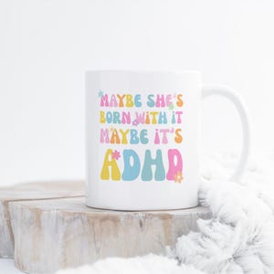 May include: White ceramic mug with the text "MAYBE SHE'S BORN WITH IT MAYBE IT'S ADHD" in colorful, retro-style lettering. The mug has a curved handle and is set against a neutral background.