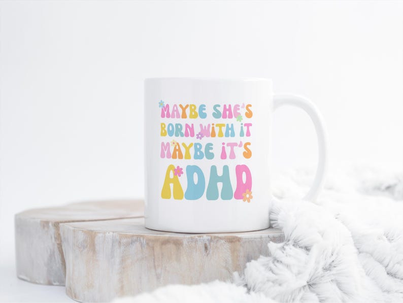 May include: White ceramic mug with the text "MAYBE SHE'S BORN WITH IT MAYBE IT'S ADHD" in colorful, retro-style lettering. The mug has a curved handle and is set against a neutral background.