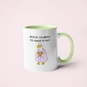 May include: White ceramic mug with a green handle and interior. The mug features a cartoon duck wearing a pink robe and holding a cup. Text on the mug reads: "Bestie, I'm about to crash tf out."