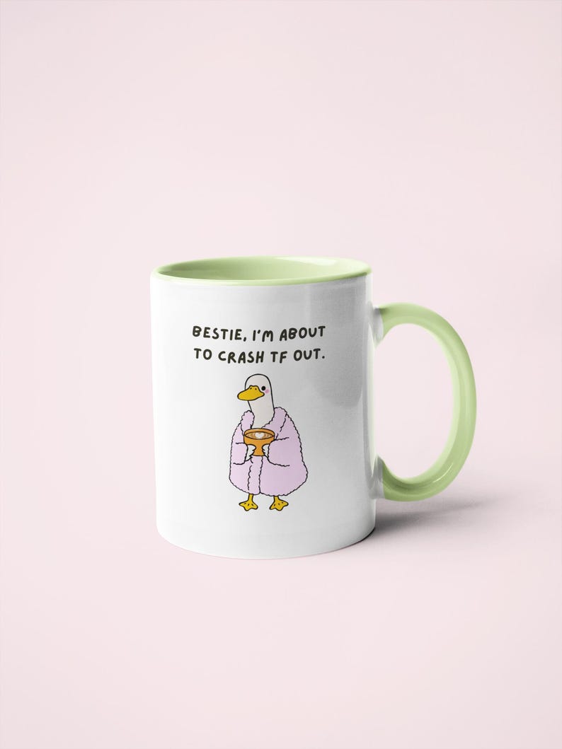 May include: White ceramic mug with a green handle and interior. The mug features a cartoon duck wearing a pink robe and holding a cup. Text on the mug reads: "Bestie, I'm about to crash tf out."