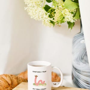 May include: White ceramic mug with a cartoon worm wearing a cowboy hat, the text reads "Sometimes I think but then I forget." The mug sits on a wooden board with a croissant and spoon.