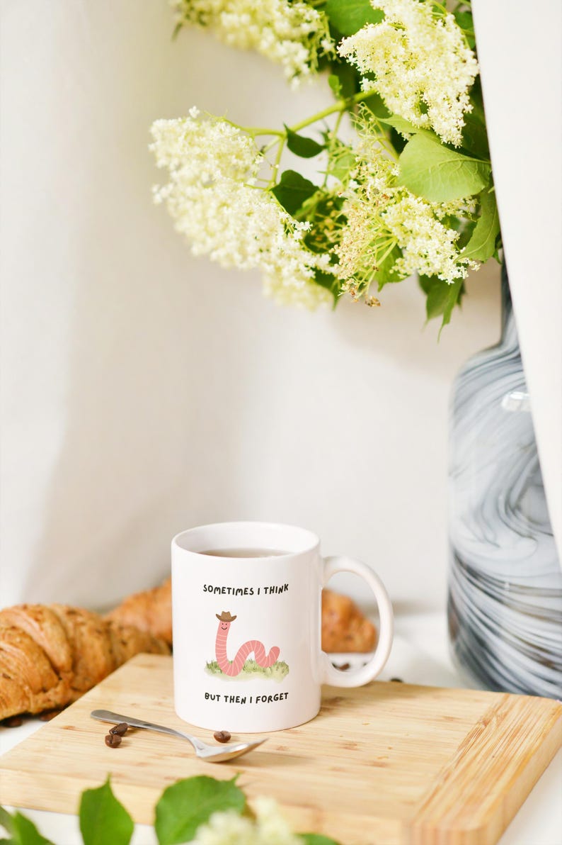 May include: White ceramic mug with a cartoon worm wearing a cowboy hat, the text reads "Sometimes I think but then I forget." The mug sits on a wooden board with a croissant and spoon.