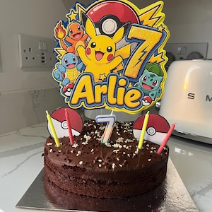Könnte beinhalten: Ein runder Schokoladen-Geburtstagskuchen mit einem großen, mehrschichtigen Pokémon-Tortentopper, der Pikachu, Glumanda, Schiggy, Bisasam, einen Pokéball, den Namen Arlie, die Zahl 7, vier Kerzen und zwei Pokéball-Ausschnitte zeigt.