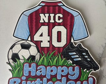 May include: A cake topper featuring a maroon and light blue soccer jersey with the name NIC and number 40, a black and white soccer ball, black soccer cleats, green grass, and the text Happy Birthday in blue glittery letters.