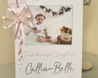 May include: A white greeting card for a 1st birthday featuring the text Happy 1st Birthday and To our beautiful Daughter Callia-Belle. The card includes a photo of a child, a pink ribbon bow, a glittery number one, and small crystals.