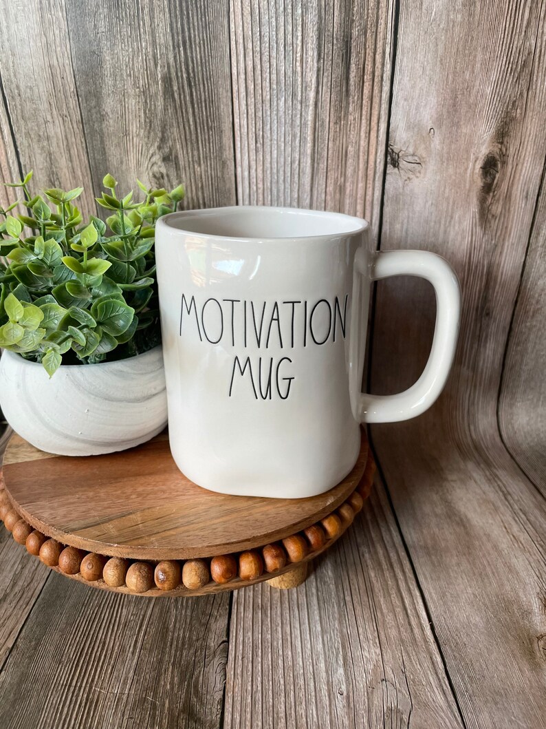 Puede incluir: Taza de cer&aacute;mica blanca con texto negro que dice "Motivation Mug". La taza est&aacute; sobre una bandeja de madera con un borde de cuentas.