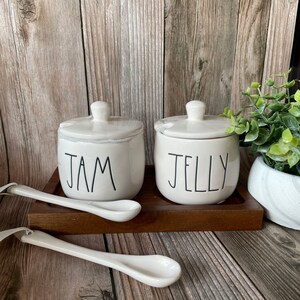 May include: Two white ceramic jars with lids, one labeled "JAM" and the other "JELLY", are displayed on a wooden tray with two white ceramic spoons.
