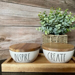 May include: Two white ceramic bowls with wooden lids, labeled "INSPIRE" and "CREATE" in black script, sit on a wooden tray. A woven basket with green foliage is in the background. The bowls and tray have a natural, rustic aesthetic.