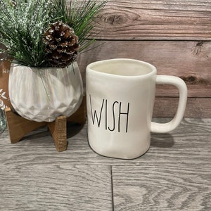 May include: White ceramic mug with a black outline of the word "WISH" on the front. The mug has a handle and is sitting on a wooden surface.