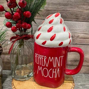 Puede incluir: Taza de cer&aacute;mica roja con un dise&ntilde;o de crema batida blanca y el texto "Peppermint Mocha". La taza est&aacute; sobre una base de madera con una rama de bayas rojas en un jarr&oacute;n de vidrio.