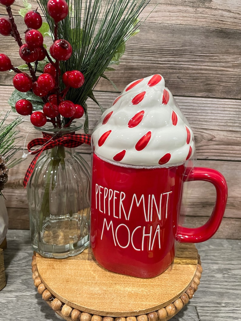 Puede incluir: Taza de cer&aacute;mica roja con un dise&ntilde;o de crema batida blanca y el texto "Peppermint Mocha". La taza est&aacute; sobre una base de madera con una rama de bayas rojas en un jarr&oacute;n de vidrio.