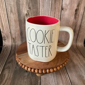 Puede incluir: Taza de cer&aacute;mica blanca con interior rojo y texto negro que dice "Cookie Taster". La taza est&aacute; sobre un posavasos de madera con borde de cuentas.