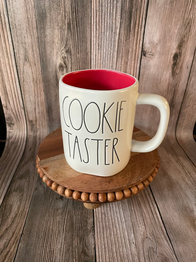 Puede incluir: Taza de cer&aacute;mica blanca con interior rojo y texto negro que dice "Cookie Taster". La taza est&aacute; sobre un posavasos de madera con borde de cuentas.
