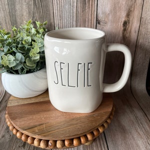 May include: White ceramic mug with a black "SELFIE" inscription. The mug is sitting on a wooden coaster with a beaded edge.