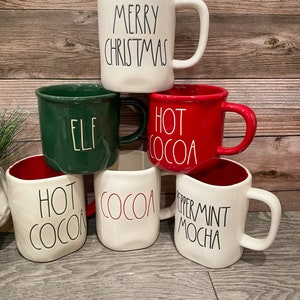 Puede incluir: Cinco tazas de cer&aacute;mica con diferentes lemas navide&ntilde;os. Las tazas son blancas con detalles rojos y verdes. Los lemas en las tazas son "Merry Christmas", "Elf", "Hot Cocoa", "Hot Cocoa" y "Peppermint Mocha".