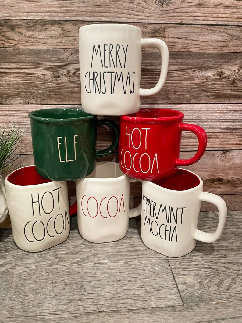 Puede incluir: Cinco tazas de cer&aacute;mica con diferentes lemas navide&ntilde;os. Las tazas son blancas con detalles rojos y verdes. Los lemas en las tazas son "Merry Christmas", "Elf", "Hot Cocoa", "Hot Cocoa" y "Peppermint Mocha".