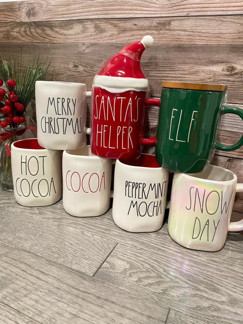 Puede incluir: Seis tazas de cer&aacute;mica blanca con diferentes frases con tem&aacute;tica navide&ntilde;a. Las tazas est&aacute;n dispuestas en forma de pir&aacute;mide sobre un suelo de madera. Las frases en las tazas son "Merry Christmas", "Santa's Helper", "Elf", "Hot Cocoa", "Cocoa", "Peppermint Mocha" y "Snow Day".